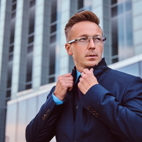 Portrait of a confident man in elegant suit and glasses correct his jacket while standing against a skyscraper background.